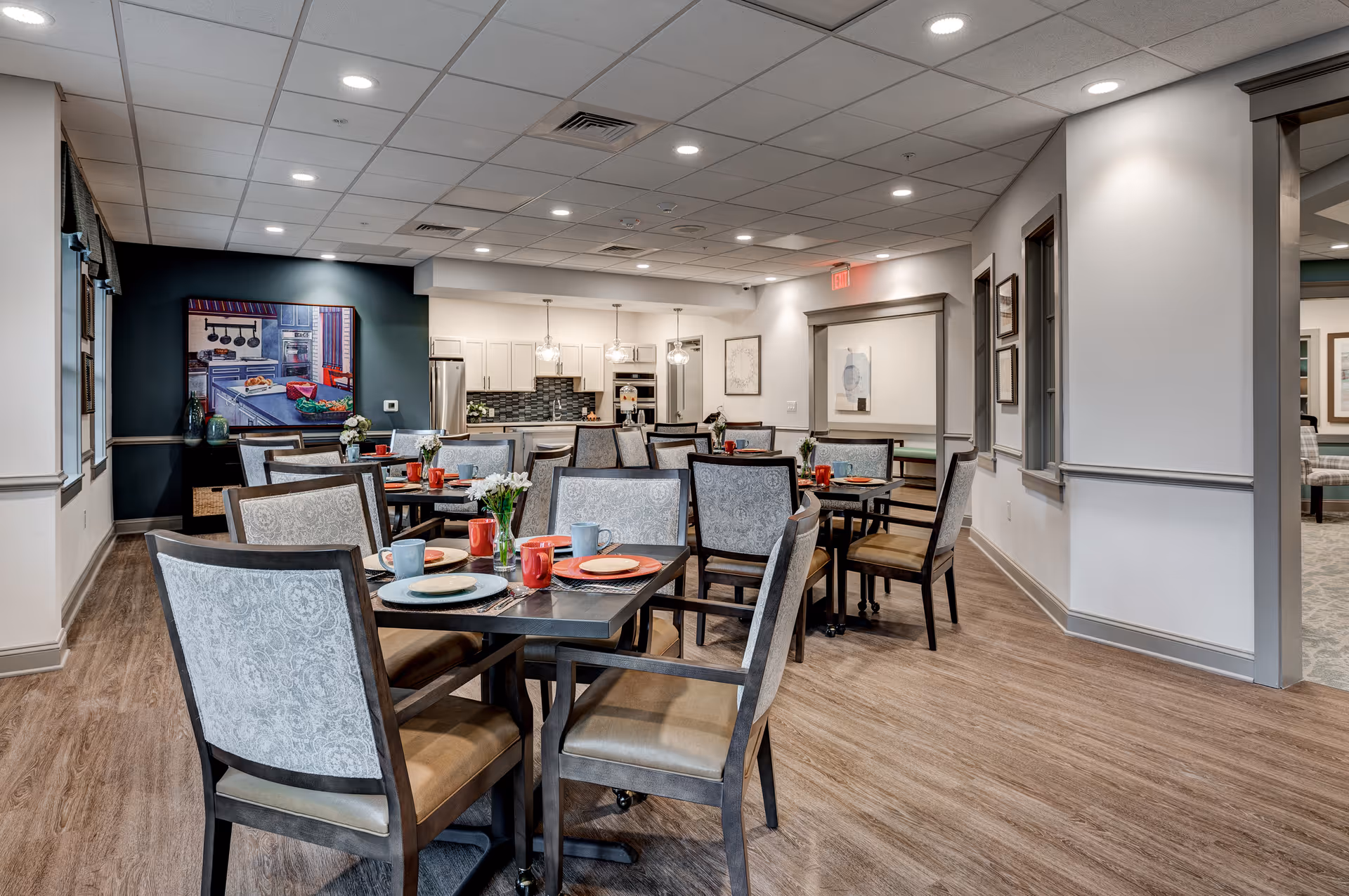 A dining room with multiple tables and chairs set with plates, cups, and utensils. The room has wood flooring, white walls with gray trim, and a ceiling with recessed lighting. In the background, there is a kitchen area with white cabinets, a stainless steel refrigerator, and pendant lights. A colorful painting of a kitchen scene is on the dark accent wall to the left.