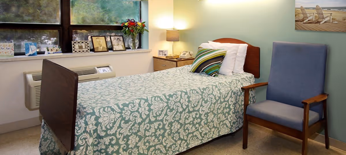 A small bedroom with a single bed covered in a green and white floral patterned bedspread. The bed has white pillows and a striped decorative pillow. Next to the bed is a wooden nightstand with a lamp, a telephone, and a framed photo. A blue upholstered wooden armchair is positioned beside the bed. The room has a large window with several framed photos and a vase of flowers on the windowsill. A picture of a beach scene with chairs is hung on the light green wall.