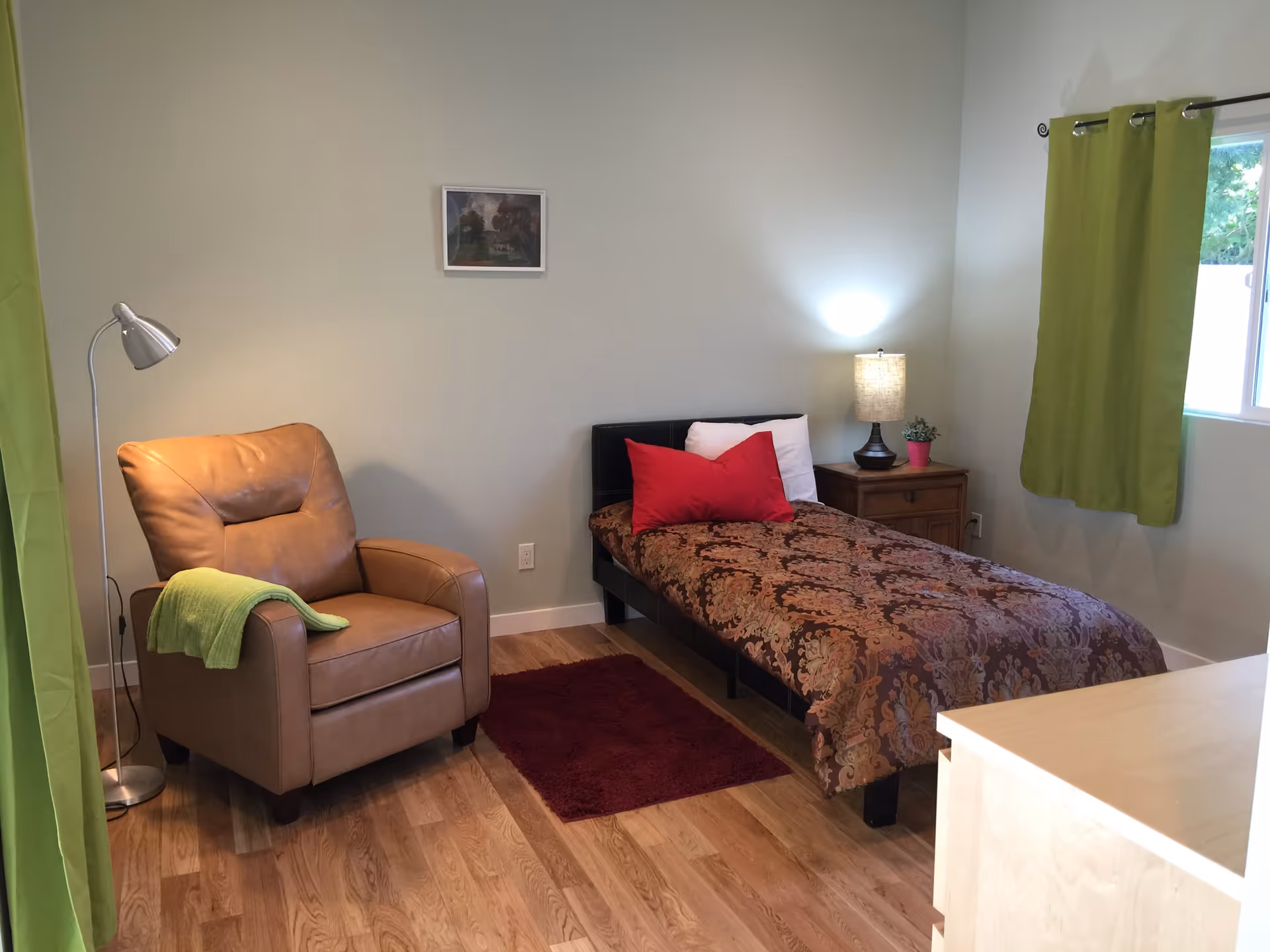 A cozy bedroom with a single bed covered in a patterned brown and orange bedspread, two red pillows, and a white pillow. Next to the bed is a wooden nightstand with a lamp and a small potted plant. A brown leather armchair with a green throw blanket is positioned near a floor lamp. The room has light-colored walls, a window with green curtains, a small red rug on the wooden floor, and a framed picture on the wall.