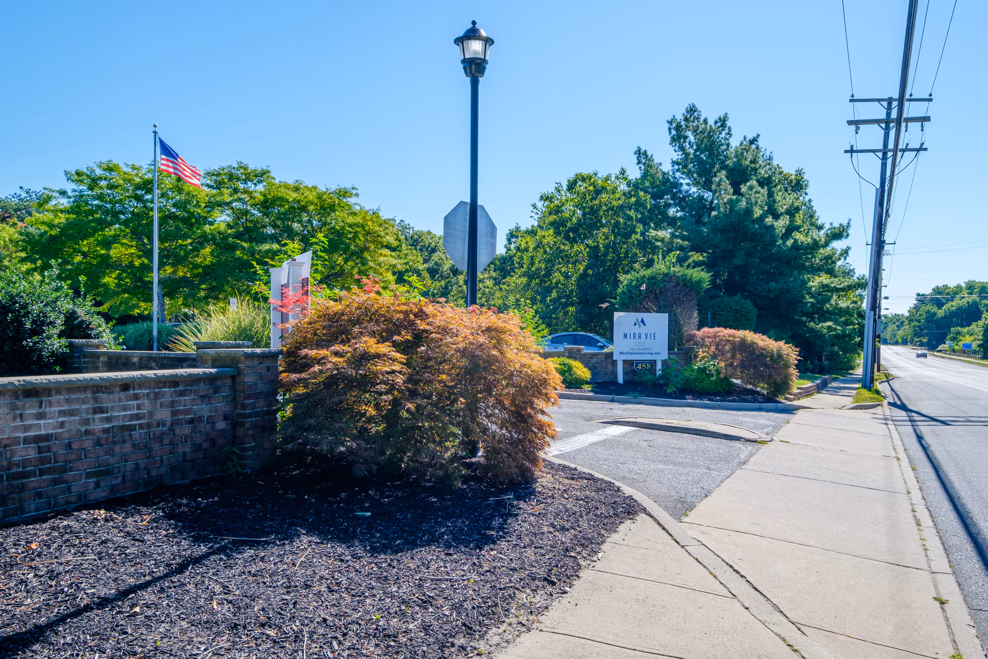 Entrance area and sign for Mira Vie at Brick assisted living along a sidewalk and roadway with landscaping and an American flag.