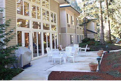 Outdoor patio area at Woodland Heights Assisted Living with white metal tables and chairs on a concrete surface, adjacent to a building with large windows. There are potted plants and trees surrounding the patio, creating a serene garden-like environment.