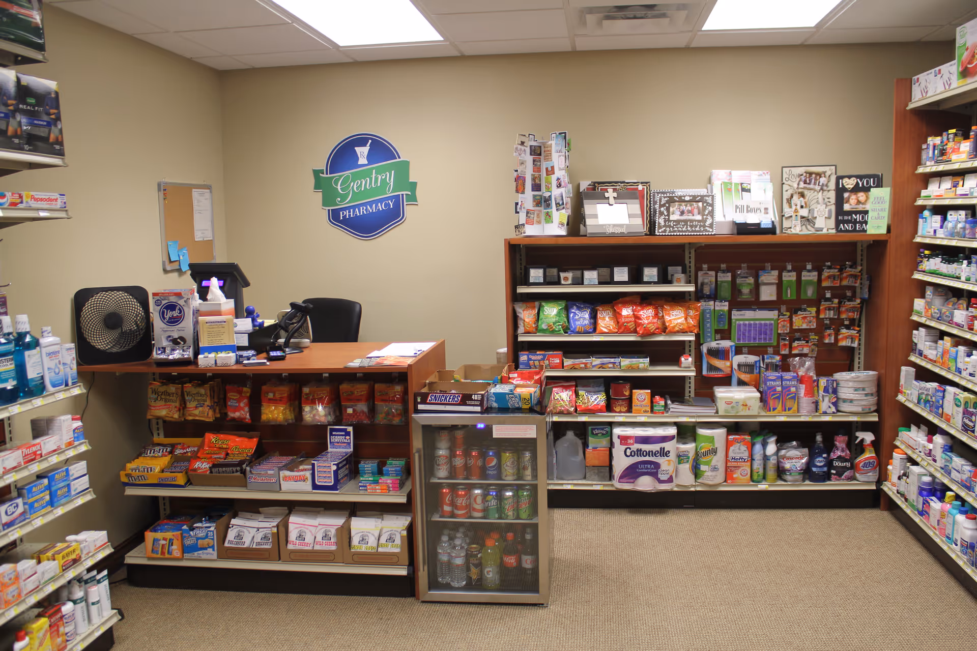 Interior view of a small pharmacy area with shelves stocked with snacks, beverages, cleaning supplies, and personal care products. A counter with a chair, a fan, and a cash register is visible. A sign on the wall reads 'Gentry Pharmacy.'