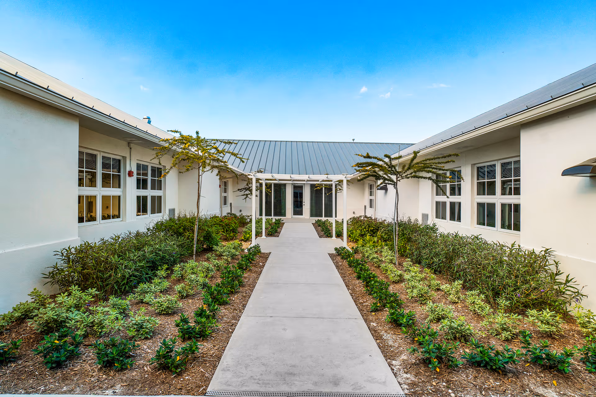 A central concrete walkway through a landscaped courtyard between two single-story white building wings under a clear blue sky.