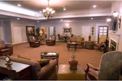 A spacious living room with multiple seating areas including armchairs and sofas arranged around coffee tables. The room features a chandelier, recessed lighting, framed artwork on the walls, and a fireplace on the right side. The carpet is patterned, and there are doors and windows visible in the background.