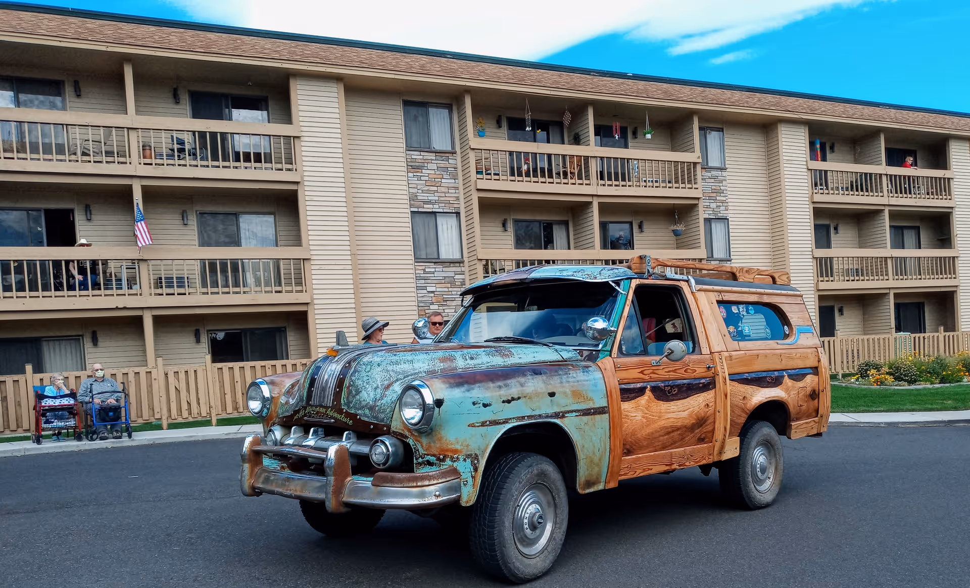 An old vintage car with a rustic, weathered look and wooden paneling is parked on a paved area in front of a multi-story assisted living community building. The building has balconies with railings and several windows. Two elderly people with walkers and two other individuals are visible near the building. The sky is partly cloudy.
