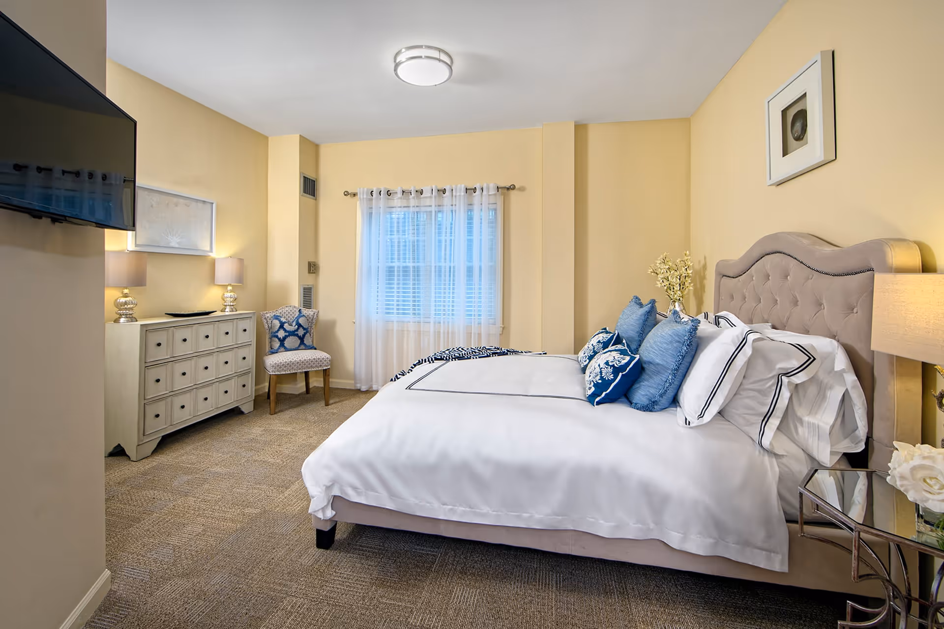 A neatly arranged bedroom with a large bed featuring white bedding and multiple blue and white pillows. The room has beige walls and carpeted floor. There is a window with sheer white curtains, a white dresser with two lamps, a patterned chair, a wall-mounted TV, and a bedside table with a lamp and flowers.