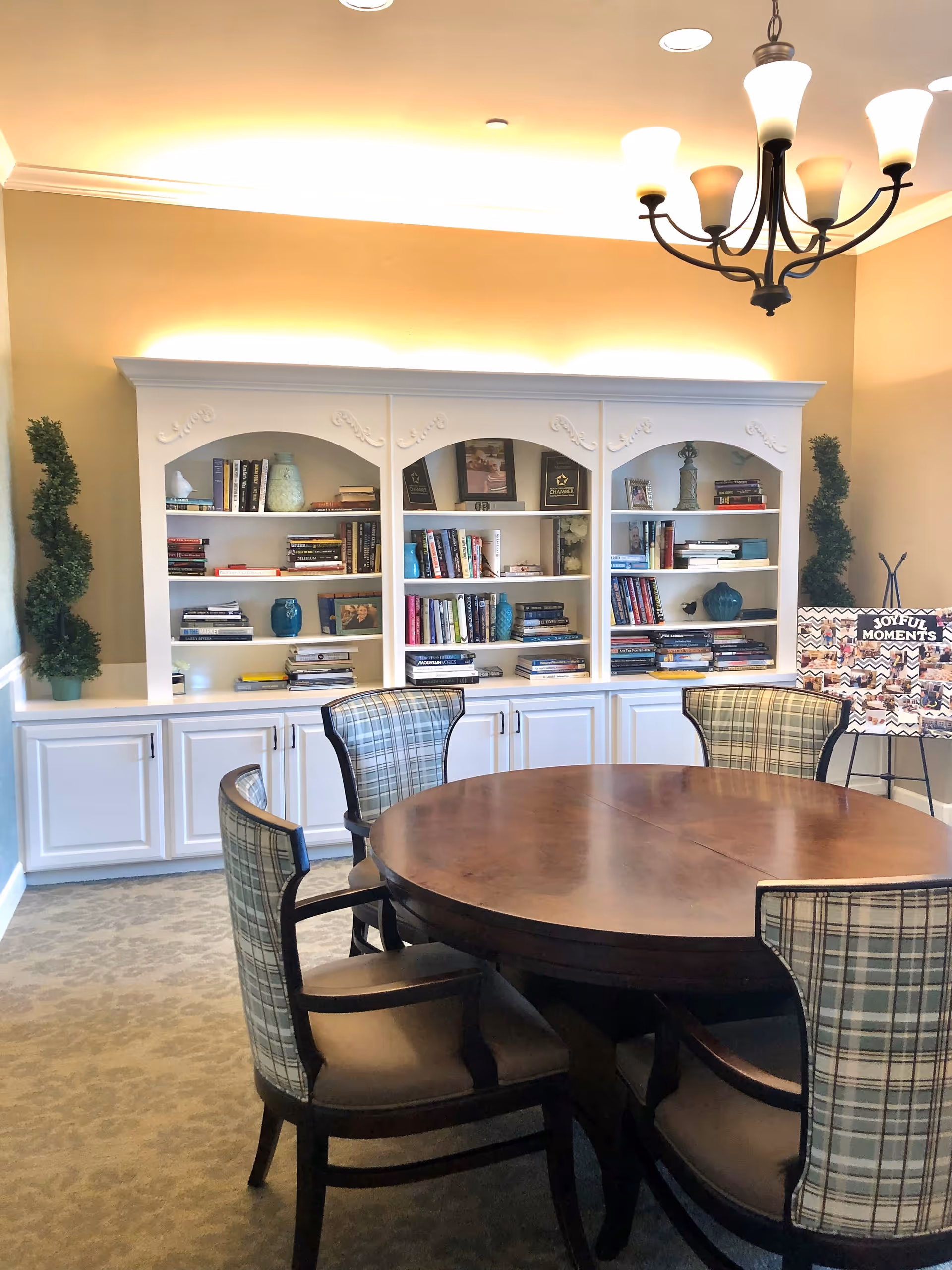 A cozy interior room with a round wooden table surrounded by four plaid upholstered chairs. Behind the table is a white built-in bookshelf filled with books, decorative items, and framed photos. Two tall green plants flank the bookshelf. A chandelier with six lights hangs from the ceiling, and a display board with the words 'Joyful Moments' is visible on the right side.