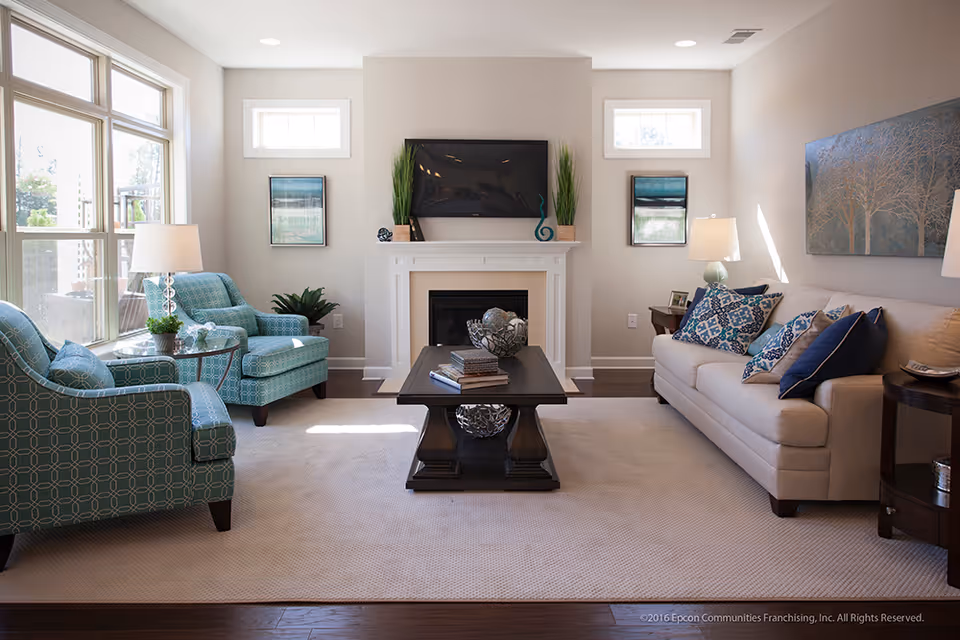 A bright and cozy living room with a beige sofa adorned with patterned blue and white pillows on the right, two teal armchairs with geometric patterns on the left, a dark wooden coffee table in the center holding decorative items and books, a white fireplace with a mounted flat-screen TV above it, and two framed artworks on the wall flanking the fireplace. Large windows on the left allow natural light to fill the room.