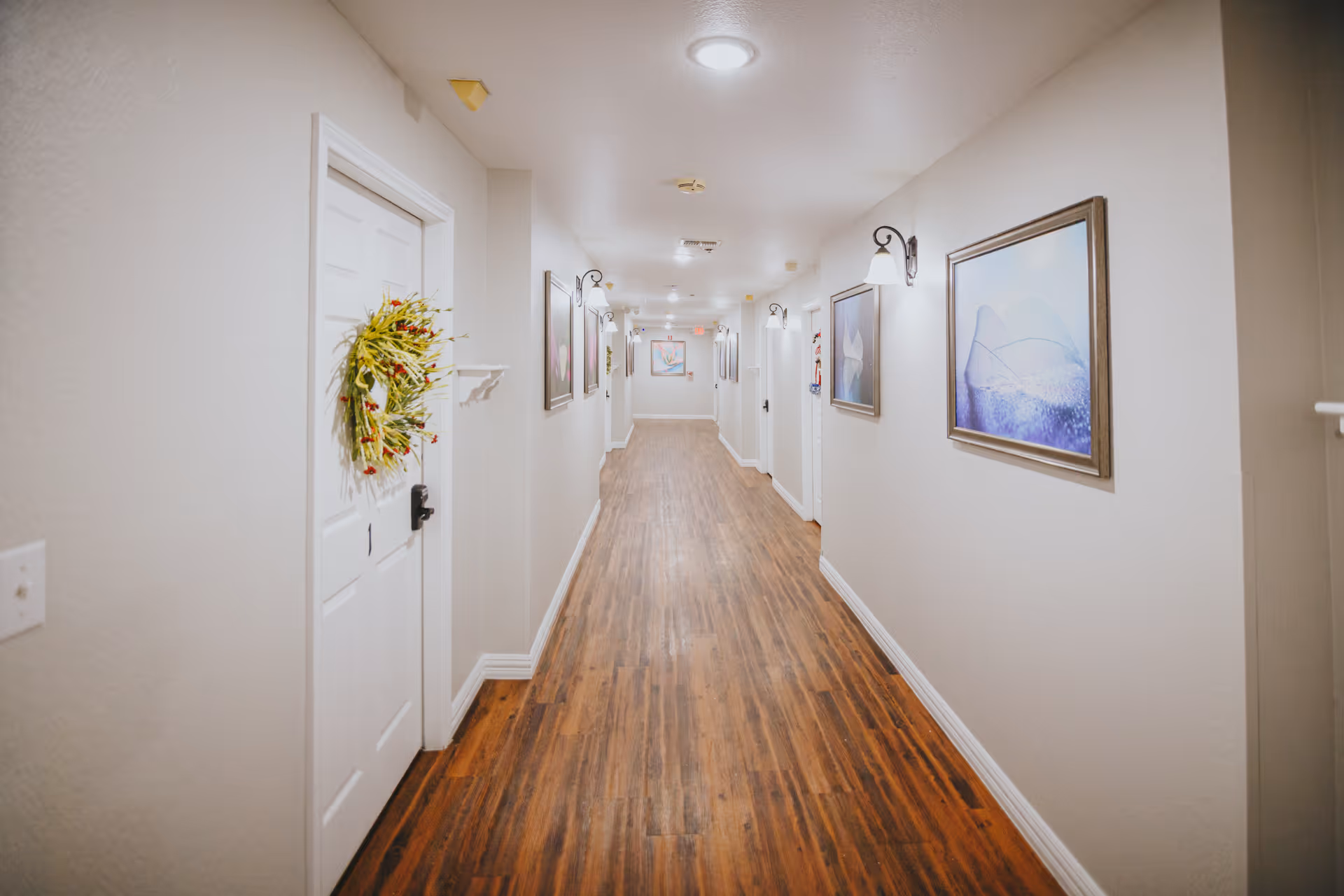 A long, well-lit hallway with wooden flooring and light-colored walls. Several white doors line the hallway, one decorated with a wreath. Wall-mounted lights and framed artwork decorate the walls.