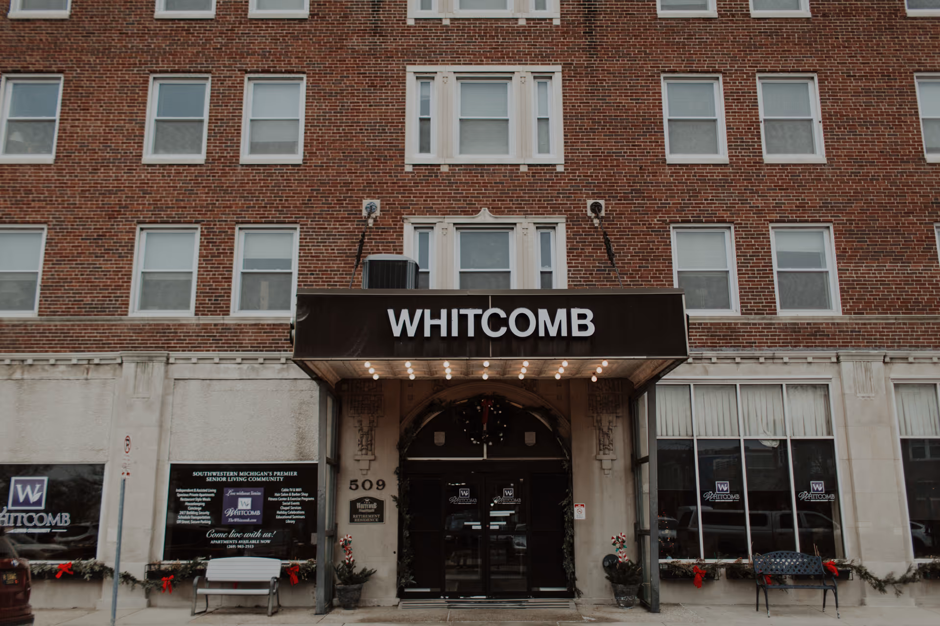 Front entrance of The Whitcomb Senior Living Tower, a multi-story brick building with a black awning displaying the name WHITCOMB above double glass doors. There are benches and holiday decorations with red bows on either side of the entrance.