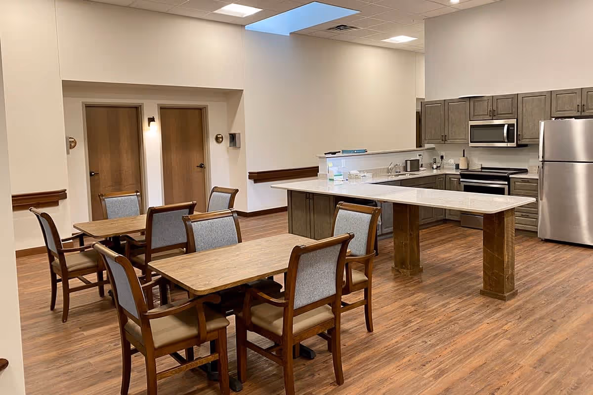 A dining area with wooden tables and upholstered chairs beside a large kitchen island and stainless steel appliances.