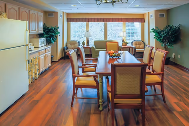 A bright and spacious dining room with a wooden dining table surrounded by eight chairs. The room features wood flooring, a kitchen area with cabinets and a refrigerator on the left, and a seating area with a sofa and two armchairs near large windows with blinds. There are two table lamps on side tables next to the sofa and green plants in the corners.