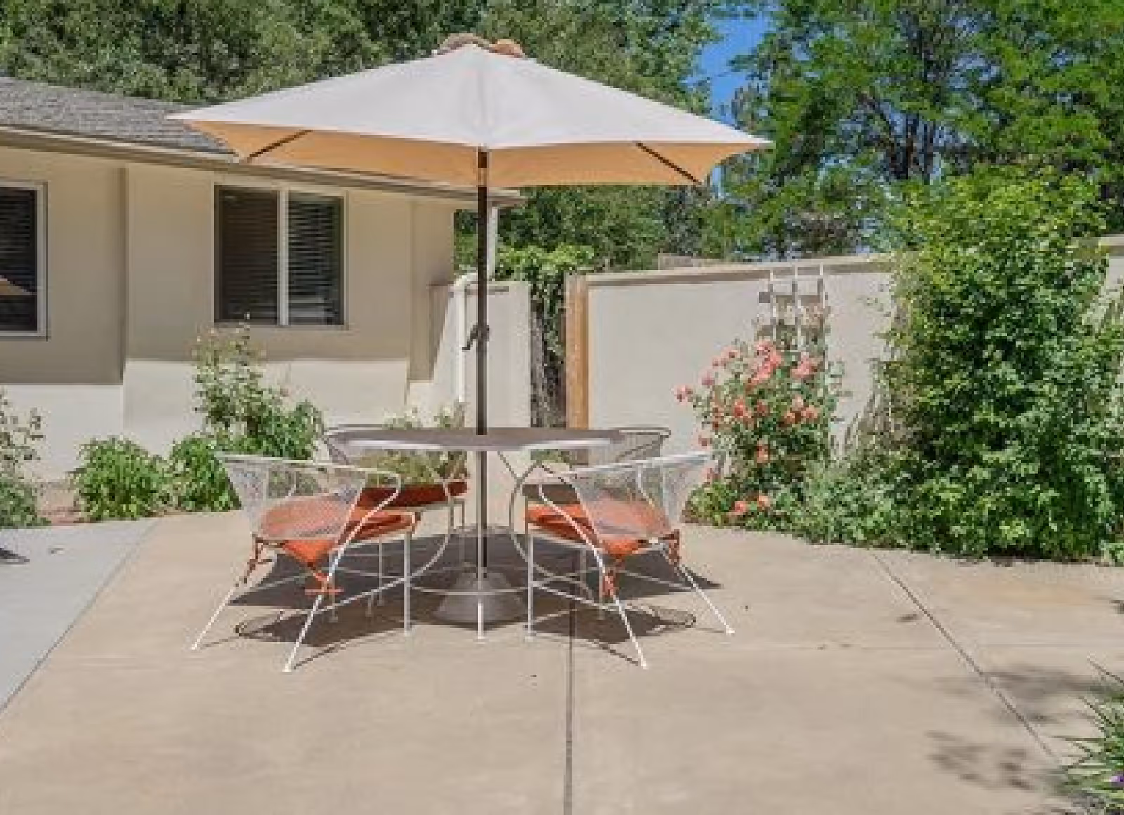 Outdoor patio with a round table, umbrella and four chairs on a concrete slab beside a single-story building and garden.