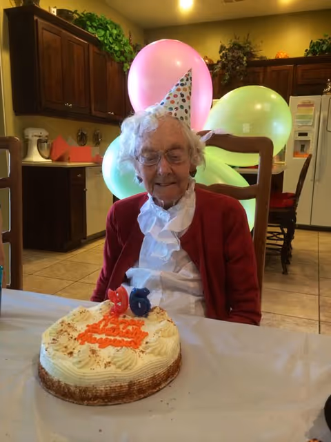 An elderly woman wearing a party hat and a red cardigan sits at a table in a kitchen with a birthday cake in front of her. The cake has candles shaped as the numbers 9 and 6. Behind her, there are colorful balloons and kitchen cabinets.