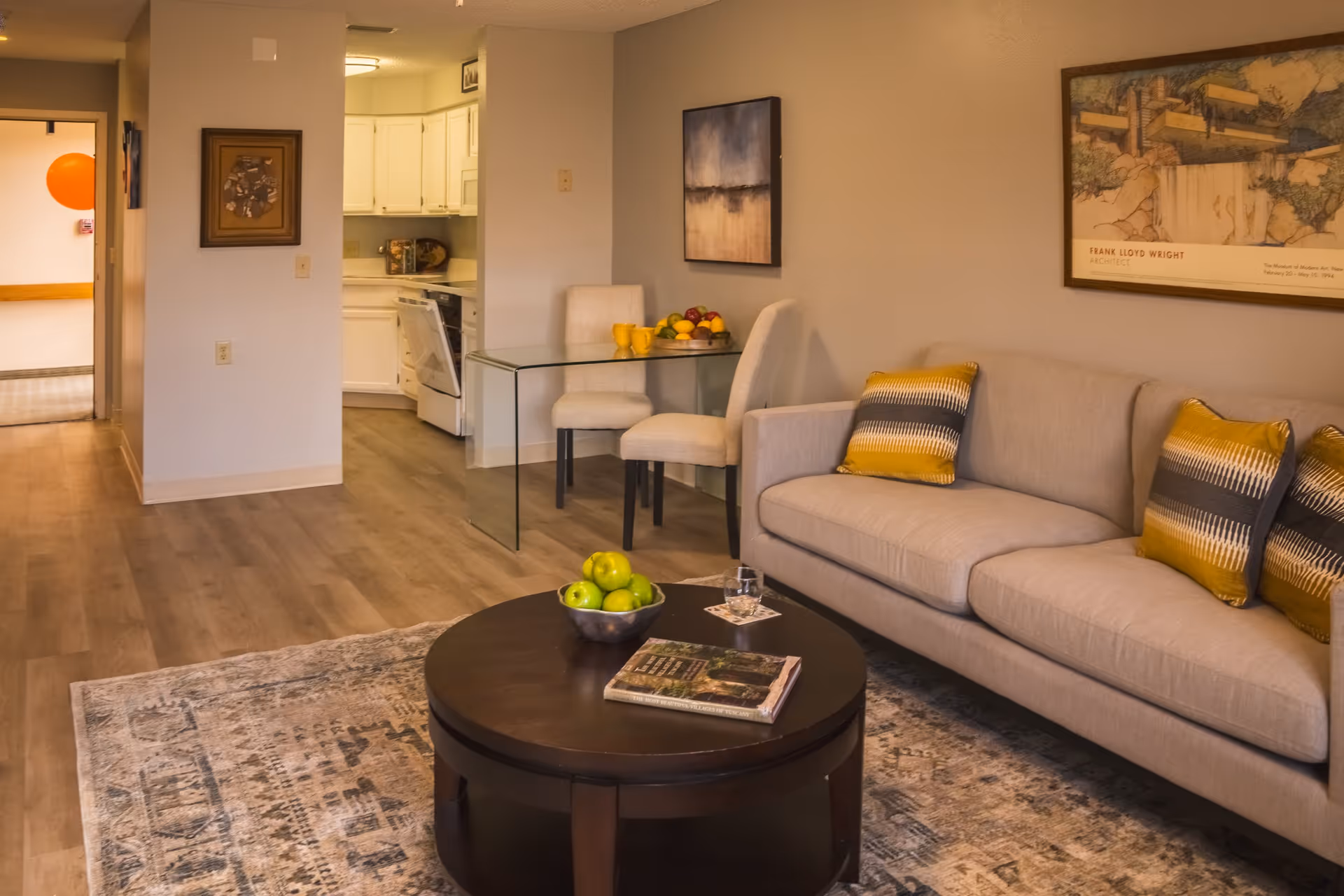 A cozy living room area with a beige sofa adorned with yellow and gray patterned pillows. In front of the sofa is a round dark wooden coffee table with a bowl of green apples, a glass of water, and a book. Behind the sofa, there is a framed artwork on the wall. To the left, a small glass dining table with two upholstered chairs is positioned near the entrance to a white kitchen. The floor is covered with light wood laminate and a patterned area rug.