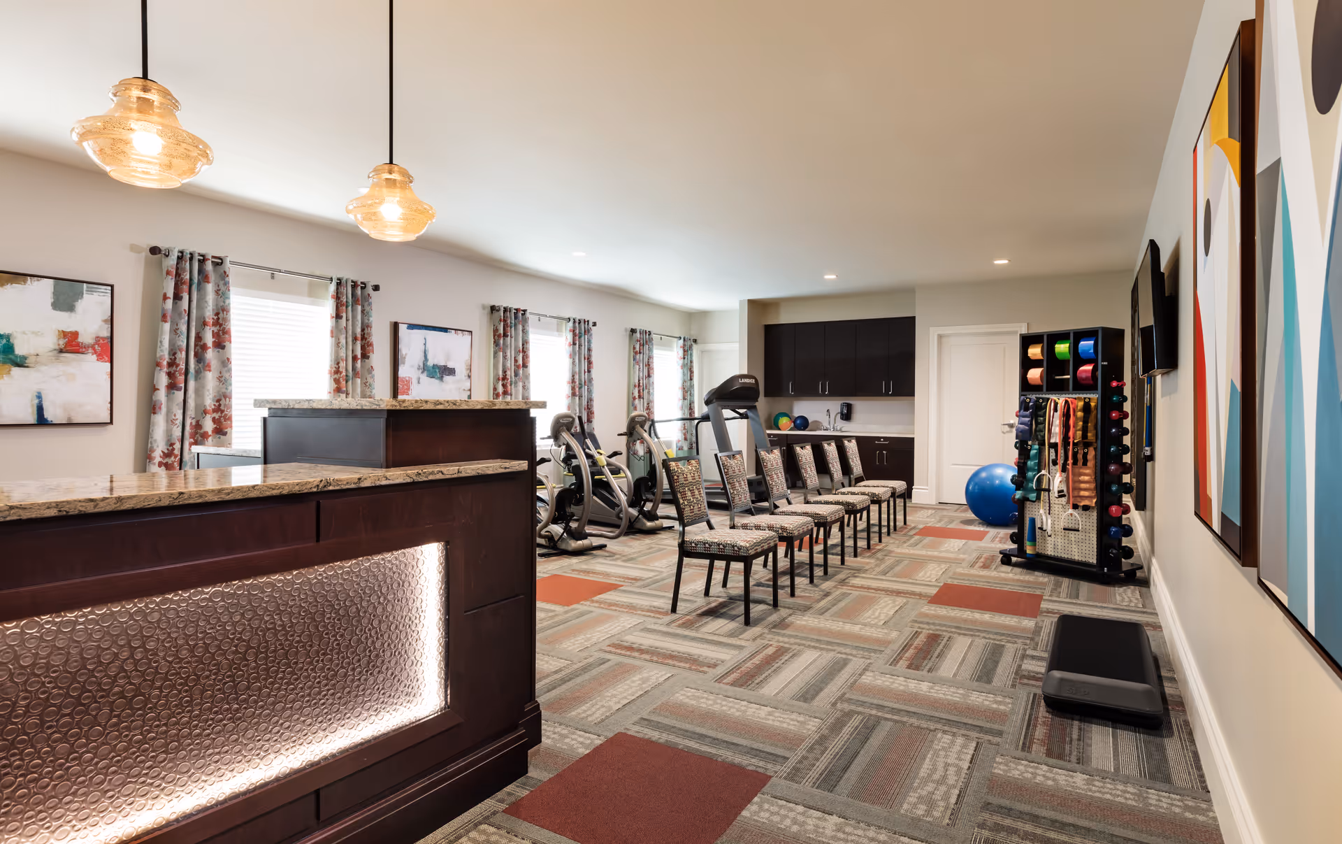A spacious fitness room with exercise equipment including stationary bikes and a treadmill, a row of patterned chairs, a rack with colorful exercise bands and weights, and modern pendant lights hanging from the ceiling. The room has large windows with floral curtains, abstract artwork on the walls, and a reception desk with a textured front panel and granite countertop.