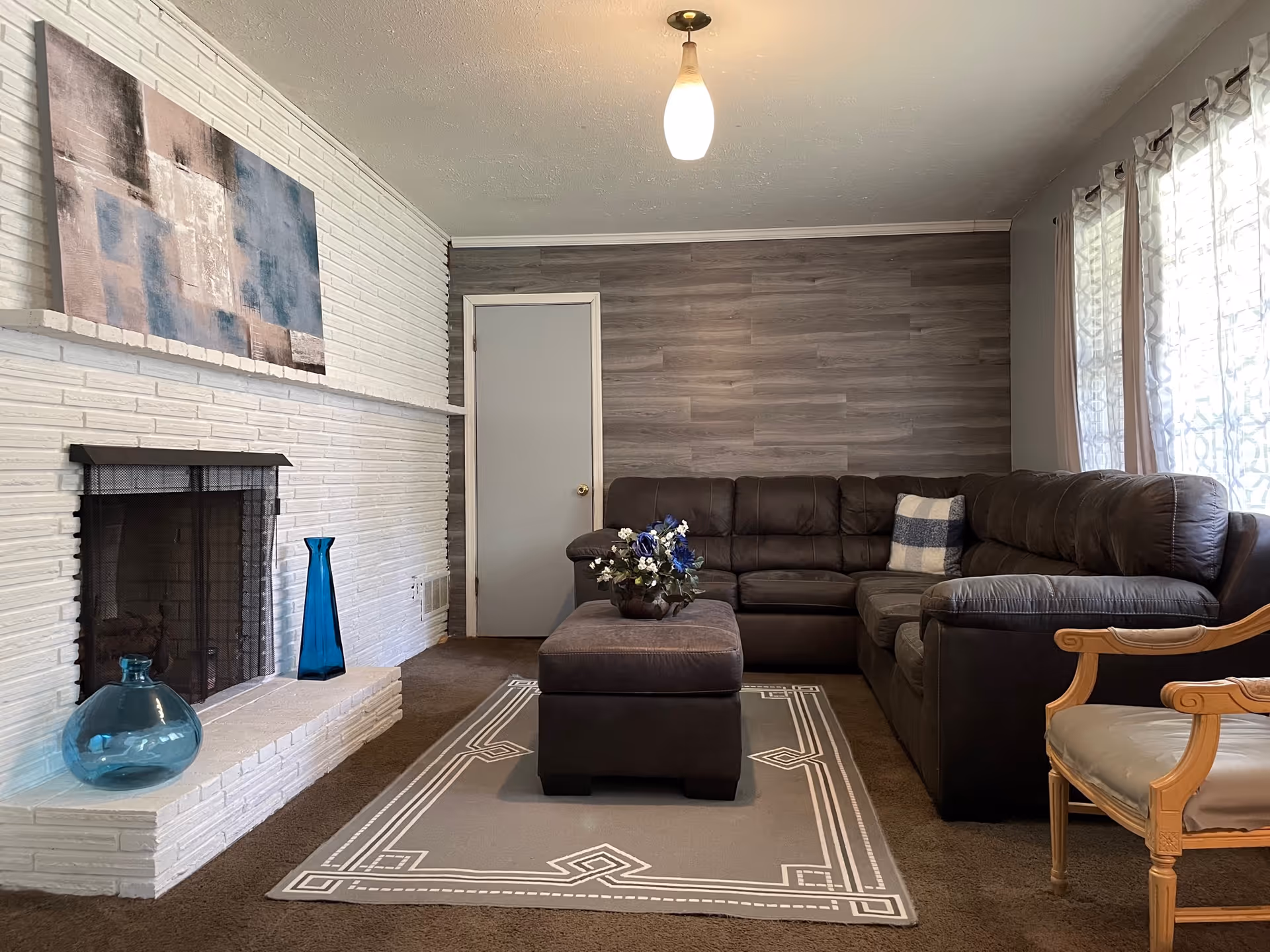 A cozy living room with a dark brown sectional sofa, a matching ottoman with a flower arrangement on top, a light gray patterned rug, a white brick fireplace with two blue vases on the hearth, a wooden armchair with a beige cushion, and a wall with wood paneling. Light filters through sheer curtains on the right side.