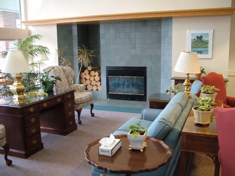 Communal living room with sofas and armchairs arranged around a tiled fireplace, side tables with lamps and potted plants, and a stack of firewood.
