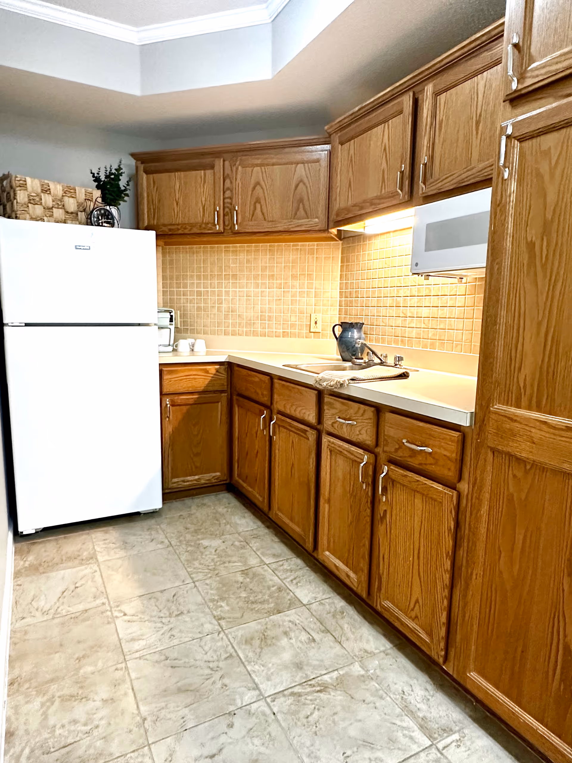 A kitchen with wooden cabinets and drawers, a white refrigerator, a microwave, and a countertop with a sink. The backsplash is tiled in a beige color, and there is a basket with greenery on top of the refrigerator. The floor is tiled in a light stone pattern.