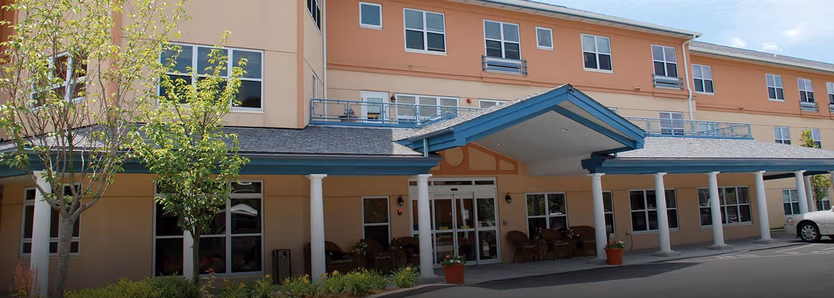 Front exterior view of a multi-story senior living facility building with a covered entrance supported by white columns, several windows, small trees, and shrubs in front, and a parked car on the right side.