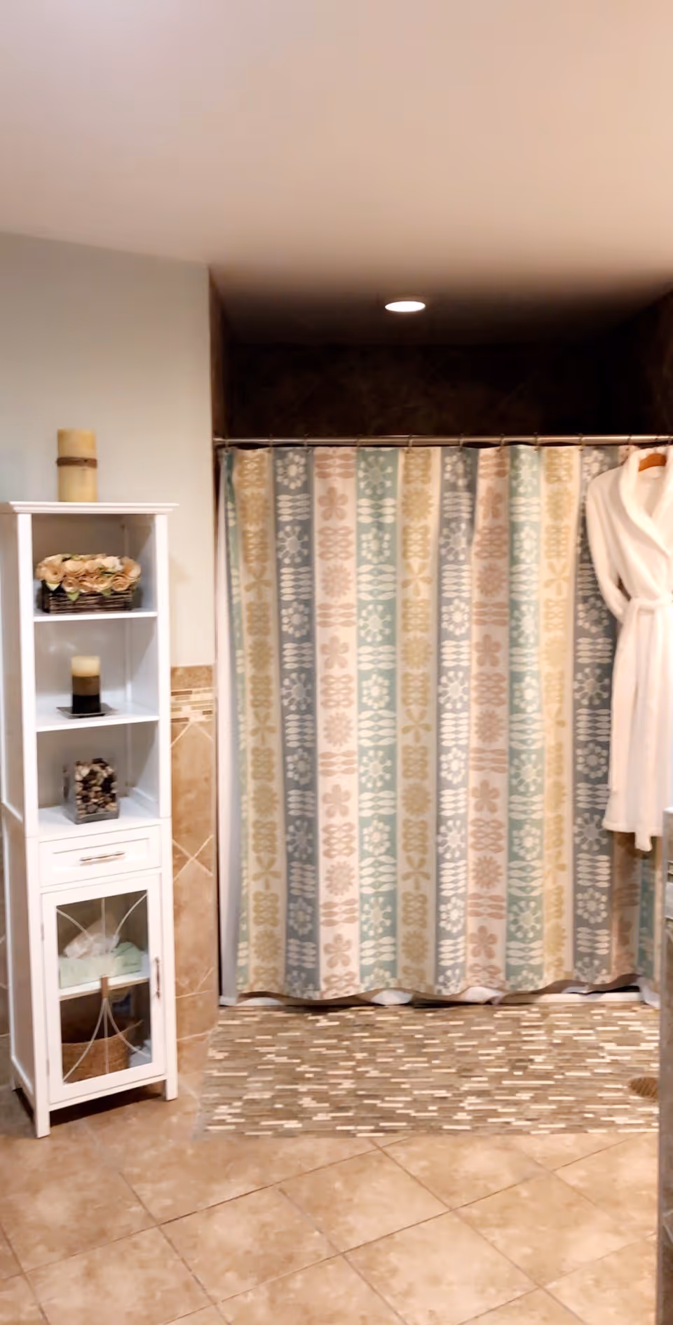 A bathroom with beige tiled floor and walls, a patterned shower curtain with vertical stripes in pastel colors, a white shelving unit holding decorative items including a candle and flowers, and a white bathrobe hanging on the right side.