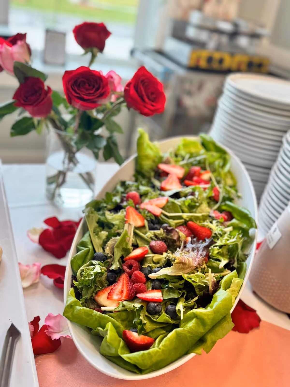 A fresh salad with mixed greens, strawberries, raspberries, and blueberries in a white oval bowl on a table. Next to the salad is a vase with red roses and rose petals scattered on the table. In the background, there are stacks of white plates and a blurred kitchen or dining area.