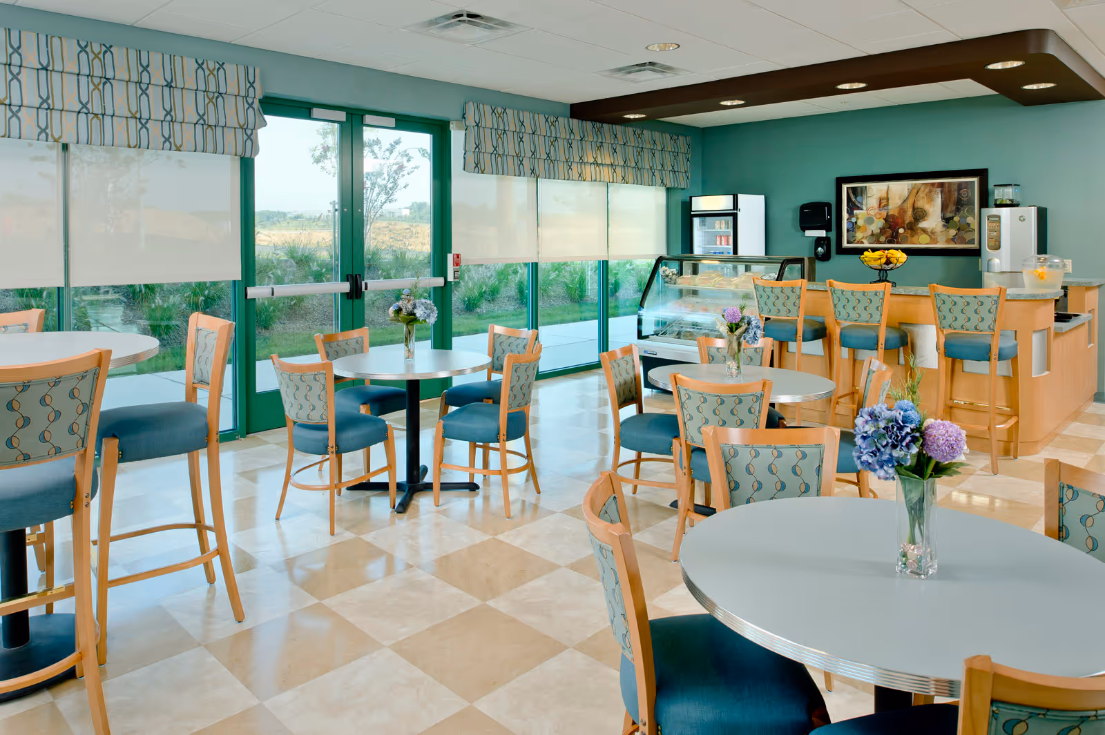 A bright and clean dining area with round tables and wooden chairs featuring blue patterned cushions. Large windows with roller shades allow natural light to fill the room. There is a counter with bar stools, a refrigerated display case, and a water dispenser. Floral arrangements are placed on the tables, and a framed artwork hangs on the teal-colored wall.