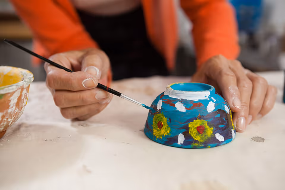 Close-up of a person painting a small ceramic bowl with a fine brush, decorating it with colorful patterns including yellow flowers and white dots on a blue background.