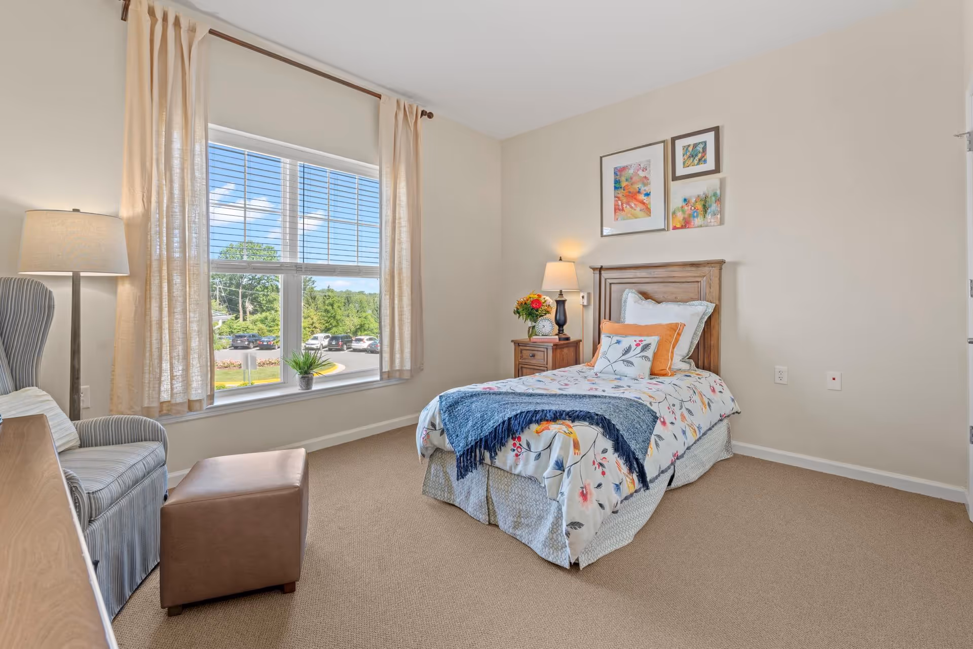 A bright and cozy bedroom with a single bed adorned with floral bedding and a blue throw blanket. There is a wooden nightstand with a lamp and a vase of flowers next to the bed. A comfortable armchair with a small ottoman is placed near a large window with beige curtains, offering a view of a parking lot and greenery outside. Three colorful framed artworks hang on the wall above the bed.