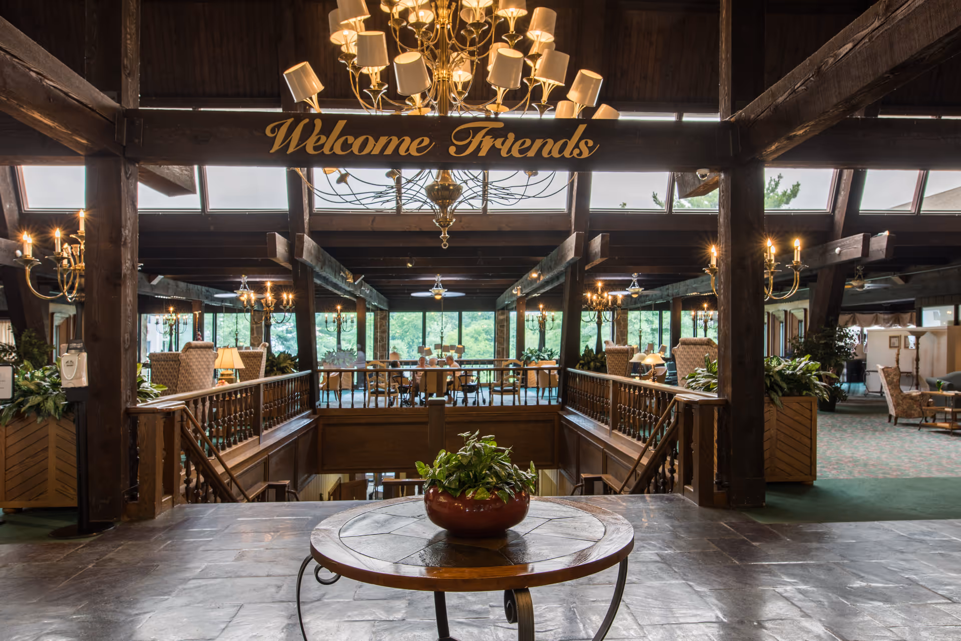 Interior view of a senior living facility lounge area with wooden beams and chandeliers. A round table with a potted plant is in the foreground. A sign above reads 'Welcome Friends'. Comfortable seating and large windows with greenery outside are visible in the background.