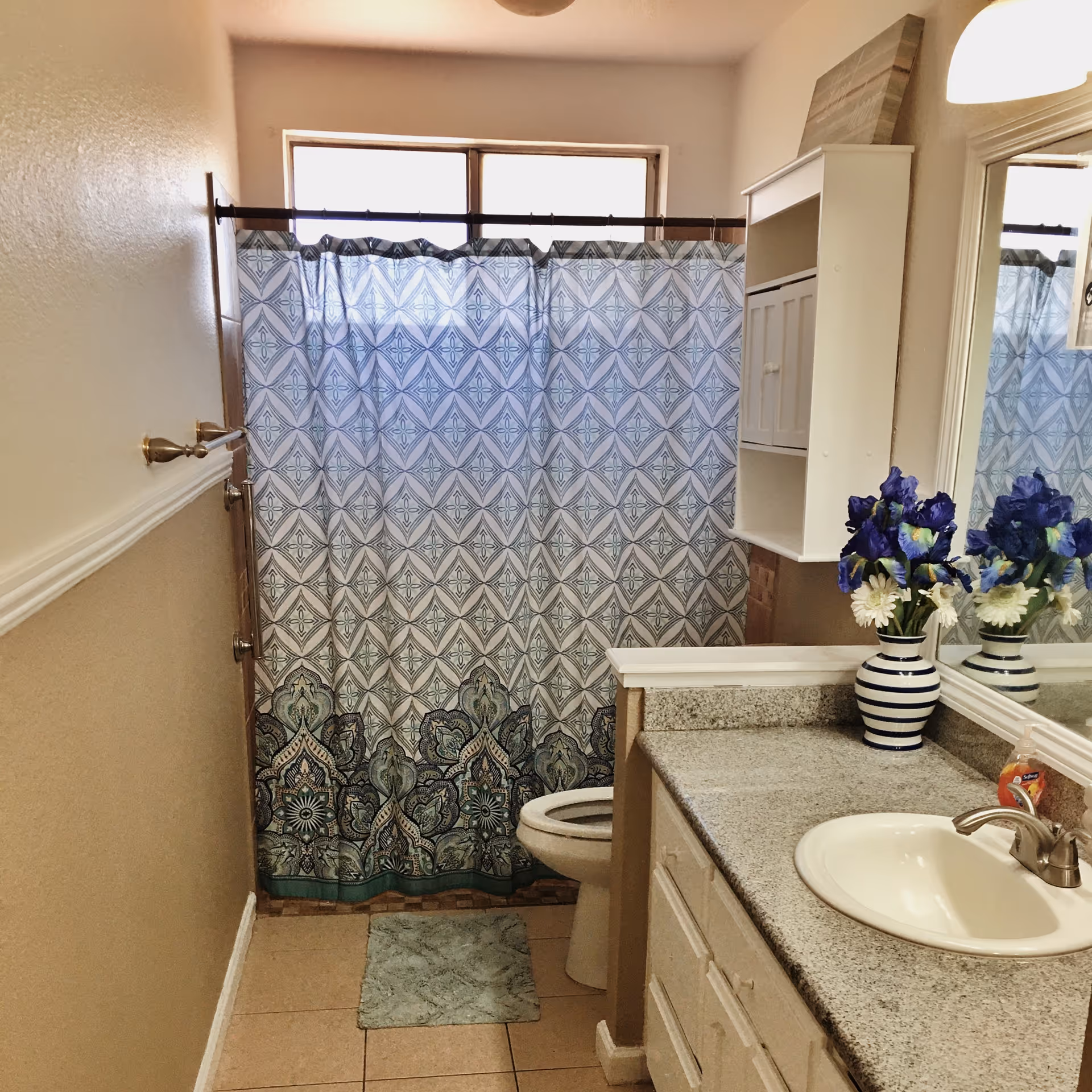 A bathroom with beige walls and tiled floor featuring a white sink with a granite countertop, a mirror above the sink, a white cabinet mounted on the wall, a toilet, and a shower with a blue and white patterned curtain. A vase with purple and white flowers is placed on the countertop.