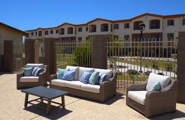 Outdoor patio with wicker sofa and chairs with cushions set before a gated courtyard and a multi-story building.