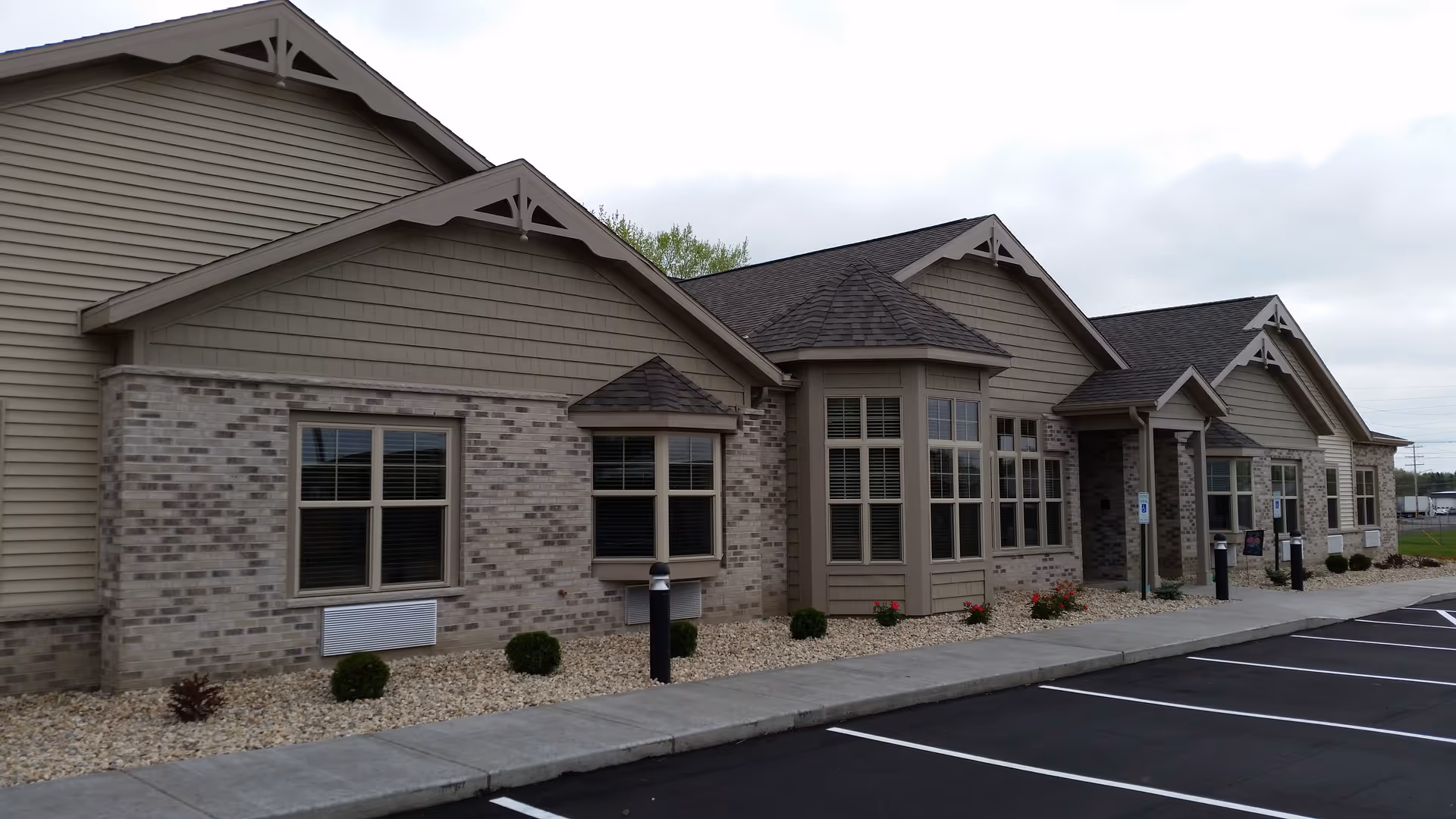 Exterior view of a single-story building with beige siding and brick facade, featuring multiple windows and a covered entrance. There is a paved parking lot with marked spaces and a sidewalk with small bushes and landscaping rocks along the building.