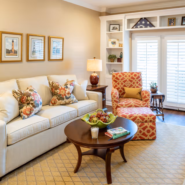 Bright living room with a cream sofa, patterned armchair, round wooden coffee table and built-in shelves by shuttered windows.