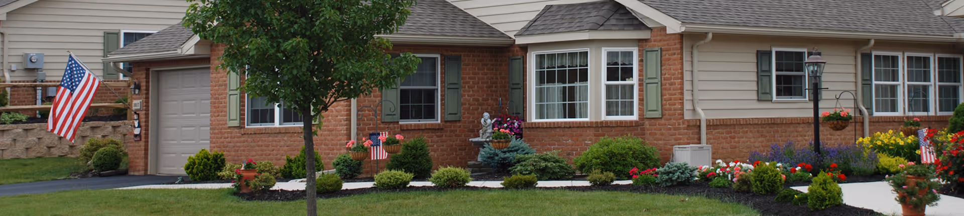 Front exterior of single-story brick and siding senior living units with garages, landscaped yards, and American flags.