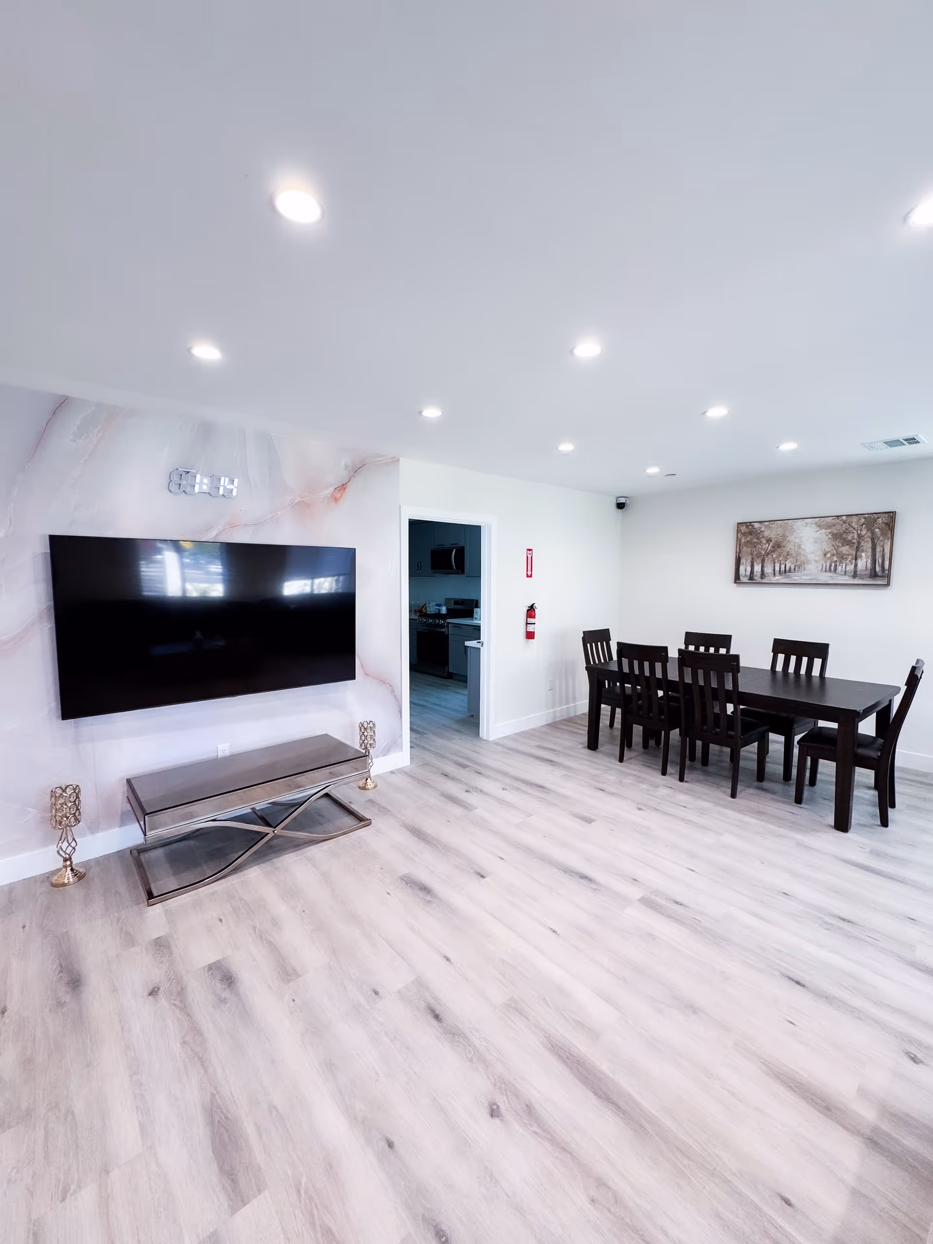 A bright and spacious room with light wood flooring and white walls. On the left wall, there is a large flat-screen TV mounted above a modern glass and metal TV stand, flanked by two decorative candle holders. To the right, there is a dark wooden dining table with six matching chairs. A framed picture of a tree-lined path hangs on the wall above the dining table. A doorway in the center leads to a kitchen area with visible appliances. The ceiling has multiple recessed lights providing ample illumination.
