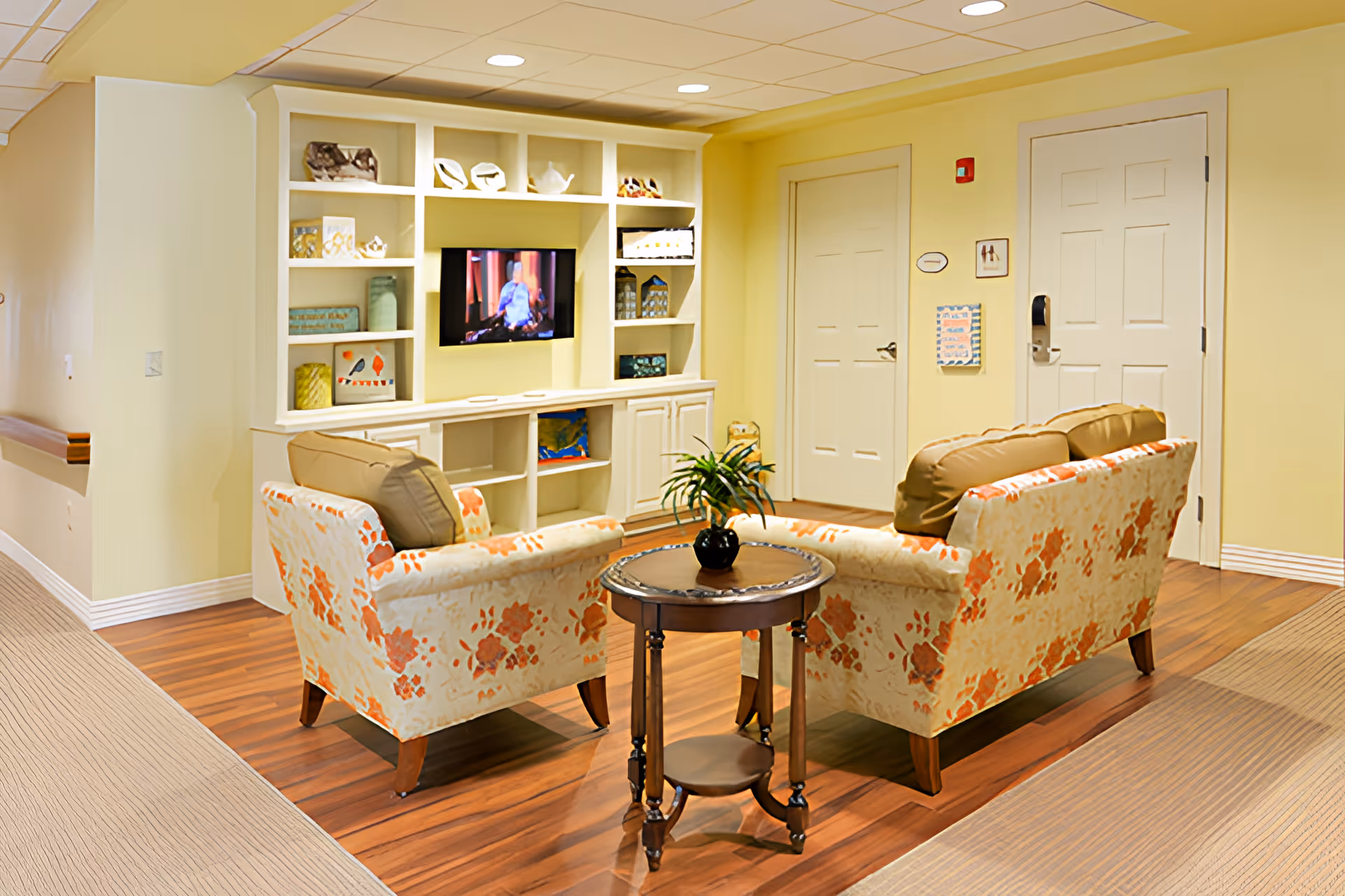 A cozy living room area in a senior living facility with two floral-patterned armchairs and a matching sofa arranged around a small round wooden table with a potted plant. A built-in white shelving unit with decorative items and a flat-screen TV is mounted on the wall. The room has light yellow walls, wood flooring, and two closed white doors in the background.