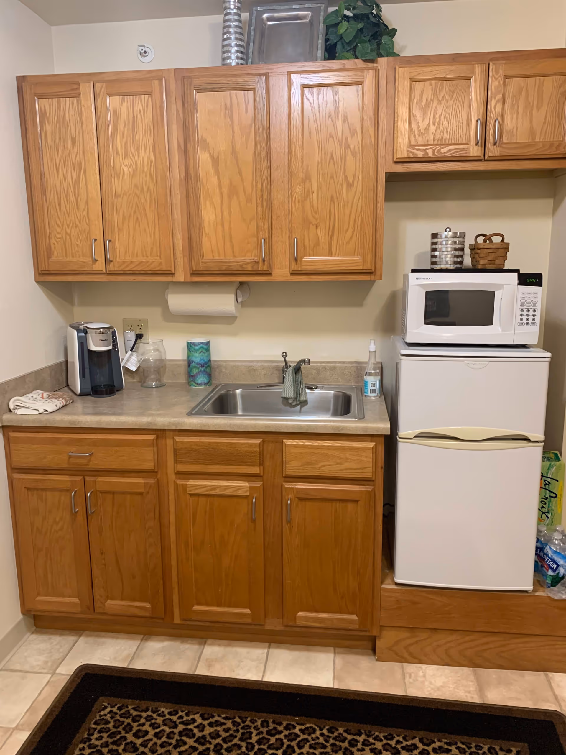 A small kitchen area with wooden cabinets above and below a countertop. On the countertop, there is a coffee maker, a glass jar, a roll of paper towels, a sink with a faucet and a cloth hanging on it, and a bottle of hand sanitizer. To the right, there is a white microwave placed on top of a white mini refrigerator. Above the microwave, there are two small containers and a small basket. The floor is tiled, and a leopard print rug is partially visible at the bottom of the image.