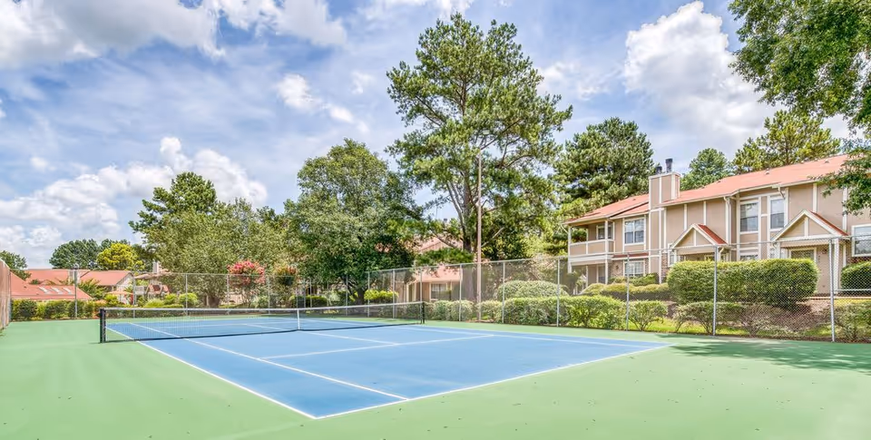 Outdoor tennis court with a blue playing surface and green surrounding area, enclosed by a chain-link fence. In the background, there are trees and residential-style buildings with red roofs under a partly cloudy sky.