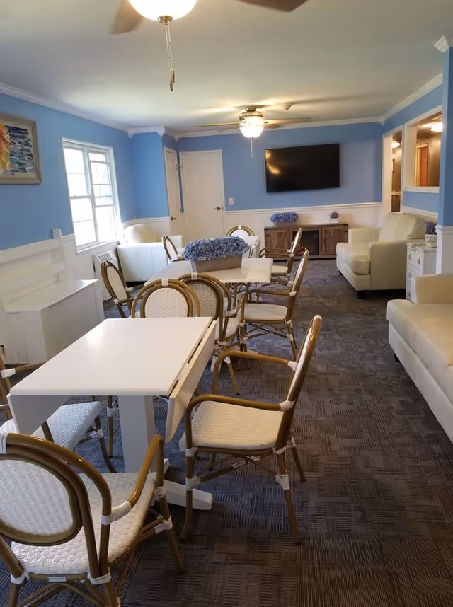 A senior living facility common area with light blue walls and carpeted floor. The room contains white tables with wicker chairs arranged around them, white sofas, a wall-mounted flat screen TV, and decorative flower arrangements on the tables and cabinet.