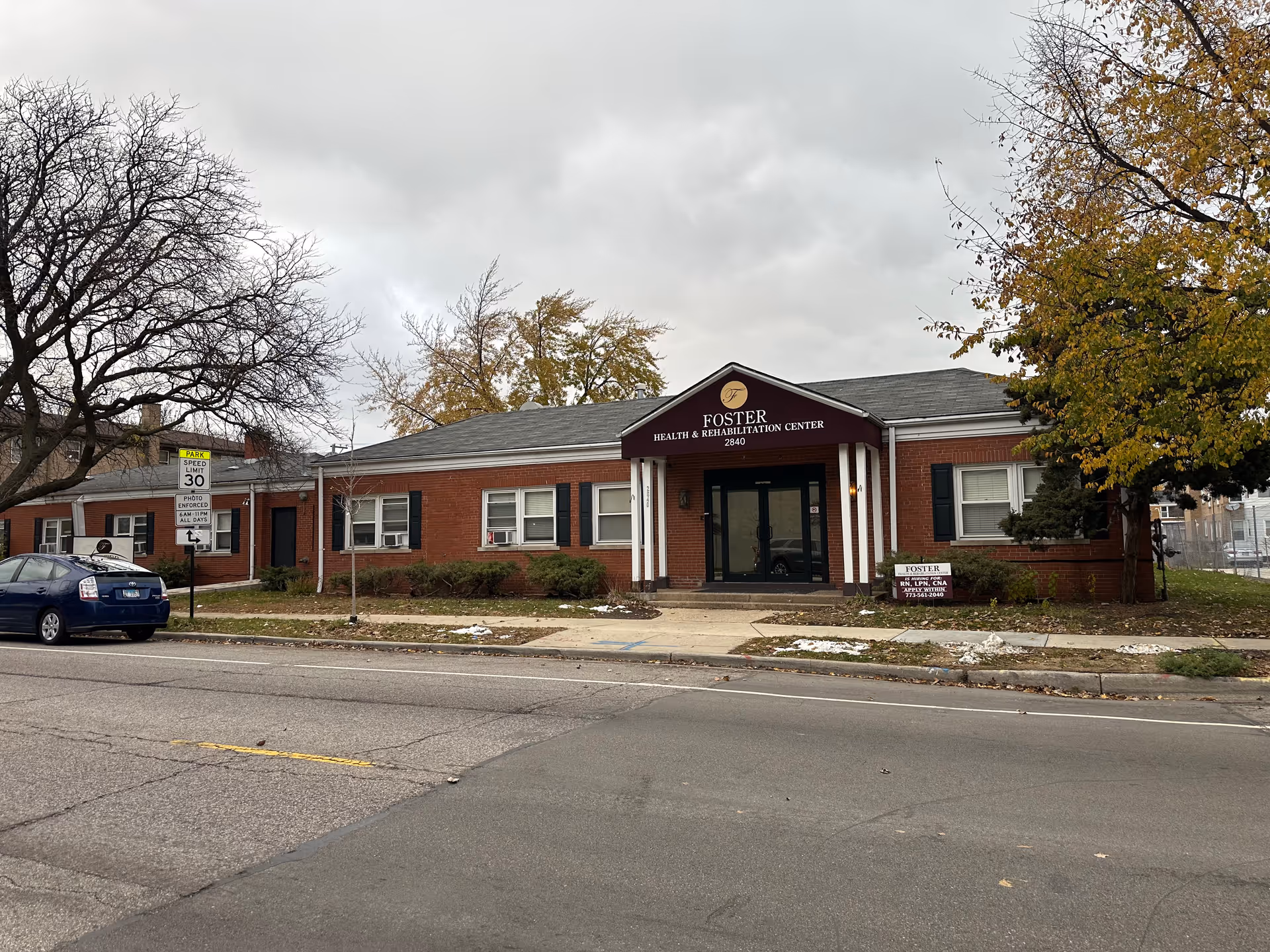 Exterior view of Foster Health & Rehabilitation Center, a single-story brick building with a maroon awning above the entrance displaying the facility name and address. There are trees with autumn leaves on either side and a parked blue car on the street in front. A speed limit sign is visible near the sidewalk.