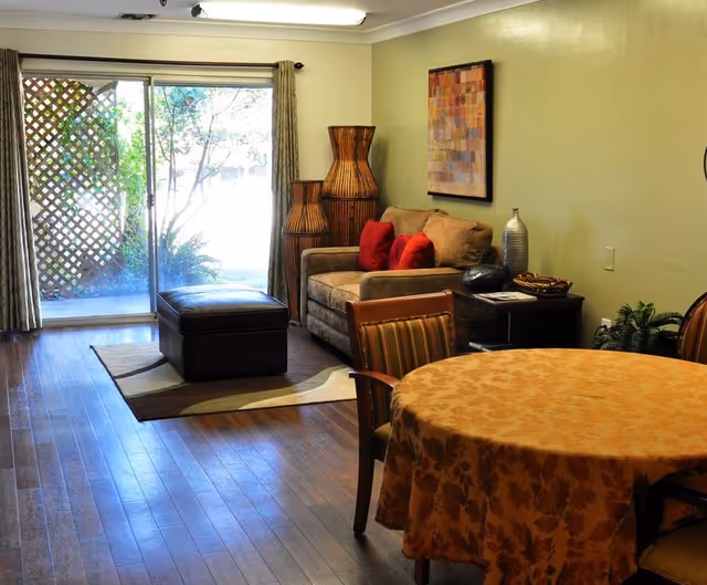 A cozy living room area with a beige loveseat adorned with red pillows, a dark ottoman on a patterned rug, and a round table covered with a floral tablecloth surrounded by chairs. There are two large decorative wicker vases in the corner and a framed abstract painting on the wall. A sliding glass door with curtains opens to an outdoor garden area.