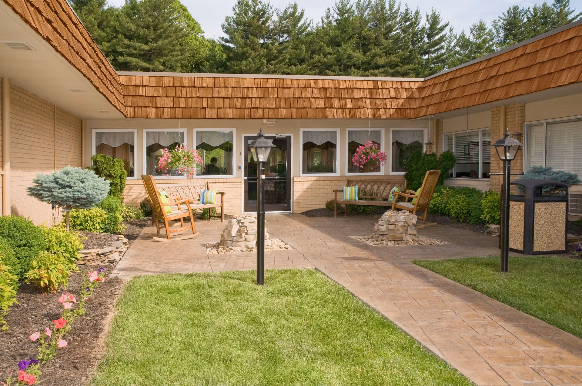 Sunny courtyard with a paved walkway leading to a building entrance, surrounded by benches, rocking chairs, hanging flower baskets and landscaping.