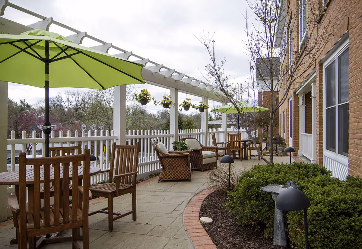Outdoor patio area at Fairmont Senior Living on Clayton with wooden tables and chairs, green umbrellas, wicker armchairs with cushions, hanging flower pots, and landscaped bushes along a brick building.