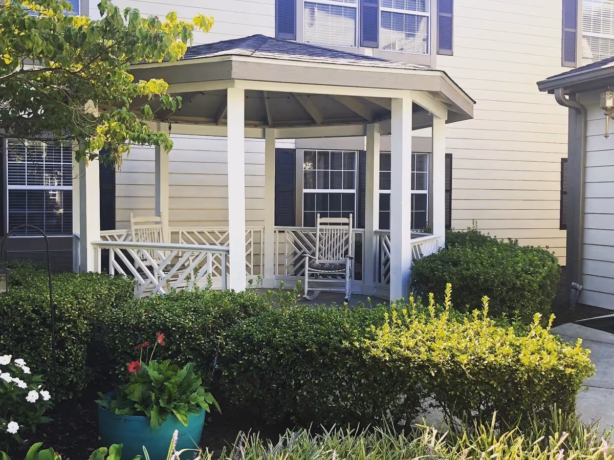 A white gazebo with a rocking chair inside, surrounded by green bushes and plants, located next to a beige building with black window shutters.