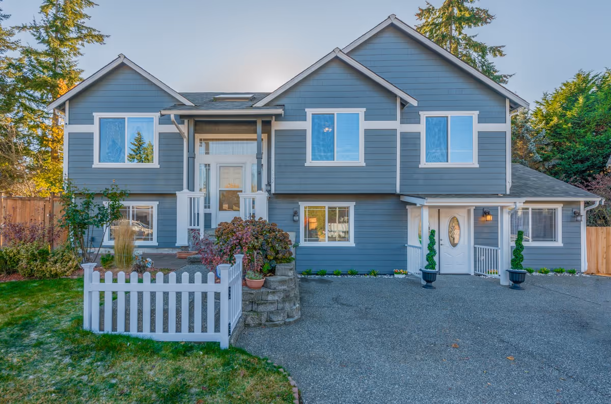 Front exterior of a two-story gray house with white trim, a driveway, small white picket fence, and landscaped yard.