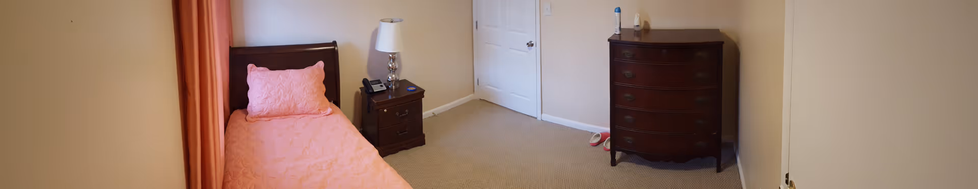A small bedroom with a single bed covered in a pink quilt and matching pillow. Next to the bed is a wooden nightstand with a silver lamp, a phone, and a small blue item. Across from the bed is a wooden chest of drawers with a bottle and a small object on top. The room has beige walls, a beige carpet, a closed white door, and pink curtains partially visible on the left side.