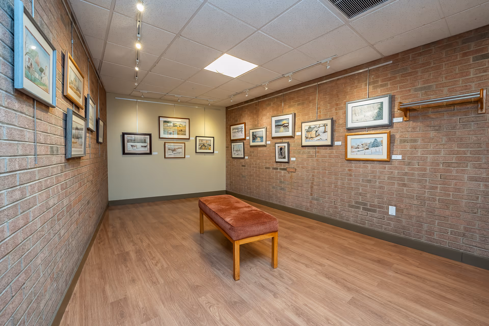 Interior view of an art gallery room with framed paintings hanging on brick and beige walls. A cushioned bench is placed in the center of the wooden floor. The ceiling has recessed lighting and a skylight.