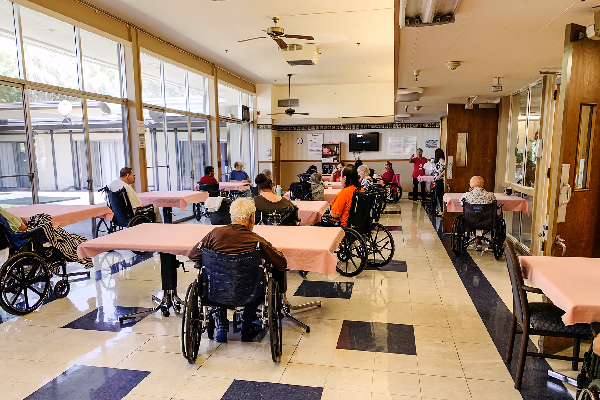 A group of elderly individuals, many in wheelchairs, seated around tables covered with pink tablecloths in a bright room with large windows. Two staff members stand near the back of the room, engaging with the residents. The room has a tiled floor with a black and white pattern and ceiling fans.