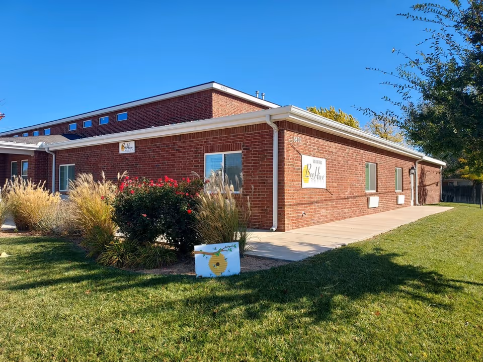 Exterior view of a single-story brick building with a sign that reads 'BeeHive Homes of Amarillo'. The building is surrounded by green grass, bushes, and ornamental grasses under a clear blue sky.